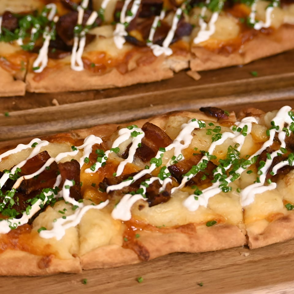 Two flatbreads topped with melted cheese, potatoes, bacon, sour cream drizzle, and chives on wooden boards.