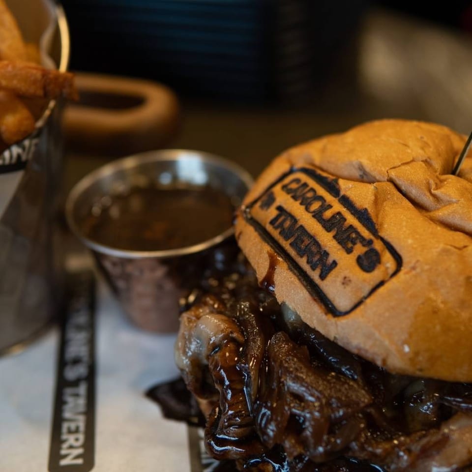 Close-up of a Caroline's Tavern burger with caramelized onions, gravy, and a side of fries.