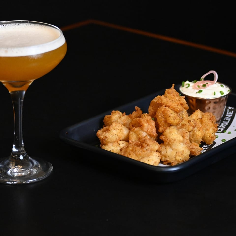 A golden cocktail with foam next to a plate of fried shrimp and dipping sauce.
