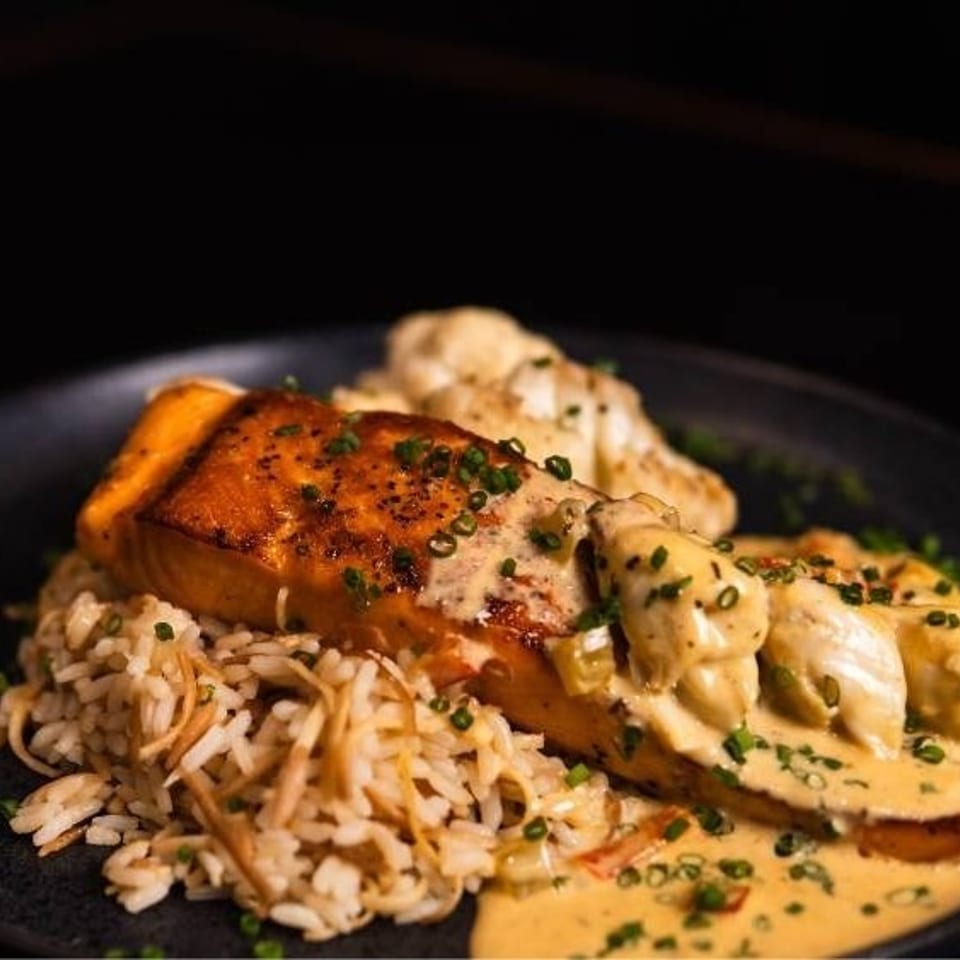 Pan-seared salmon with creamy sauce, crab meat, and rice pilaf on a dark plate.