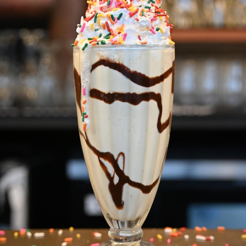 Vanilla milkshake with chocolate syrup, whipped cream, colorful sprinkles, and a cherry on top.