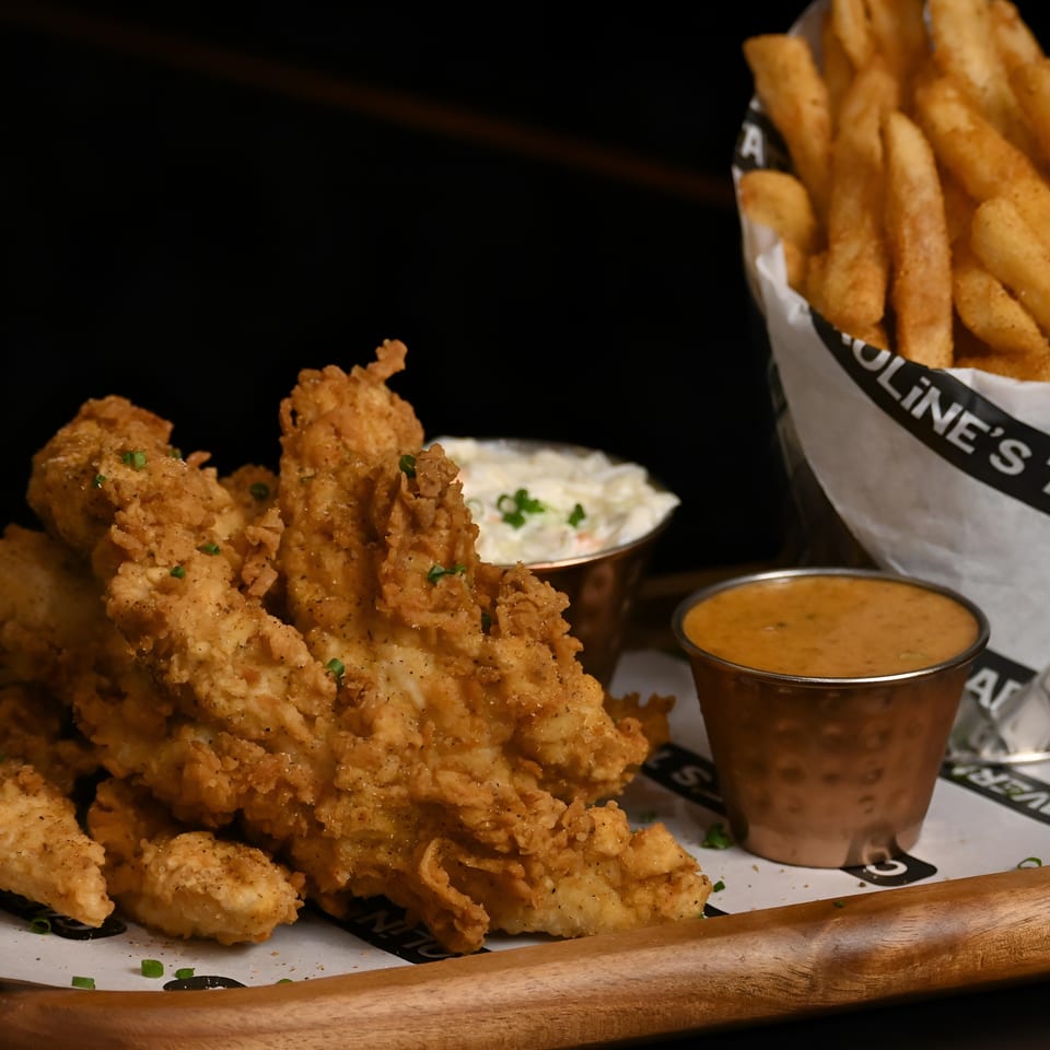 Crispy fried chicken tenders, fries in a "Caroline's Tavern" wrapper, coleslaw, and dipping sauce on a wooden tray.