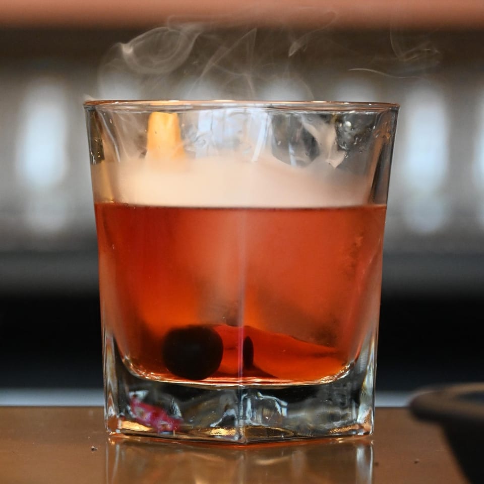 A smoky Old Fashioned cocktail with an orange peel and cherry, on a reflective bar top.