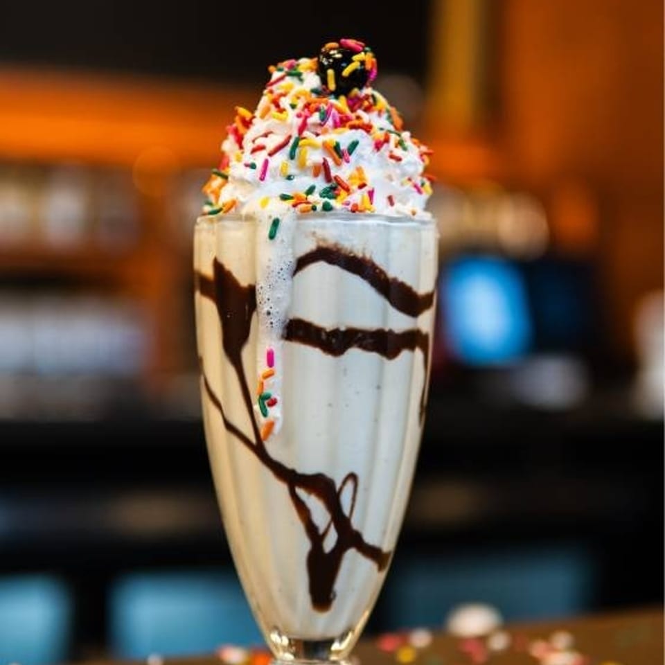 Vanilla milkshake with whipped cream, chocolate drizzle, and colorful sprinkles on a counter.