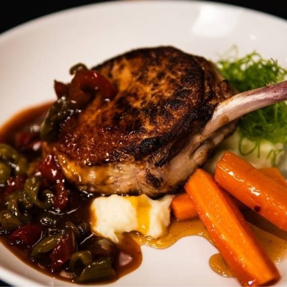 Grilled pork chop with bone, bell peppers, mashed potatoes, carrots, and gravy on a white plate.